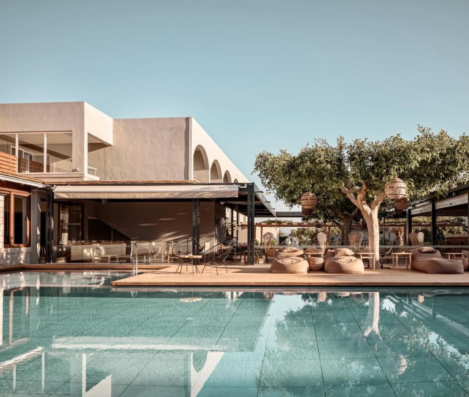 Weitläufiger Hotelpool, umgeben von modernen Gebäuden mit weißen Fassaden, einer großen Holzterrasse mit gemütlichen Sitzsäcken, Naturstühlen und schattenspendenden Bäumen mit dekorativen Lampen.