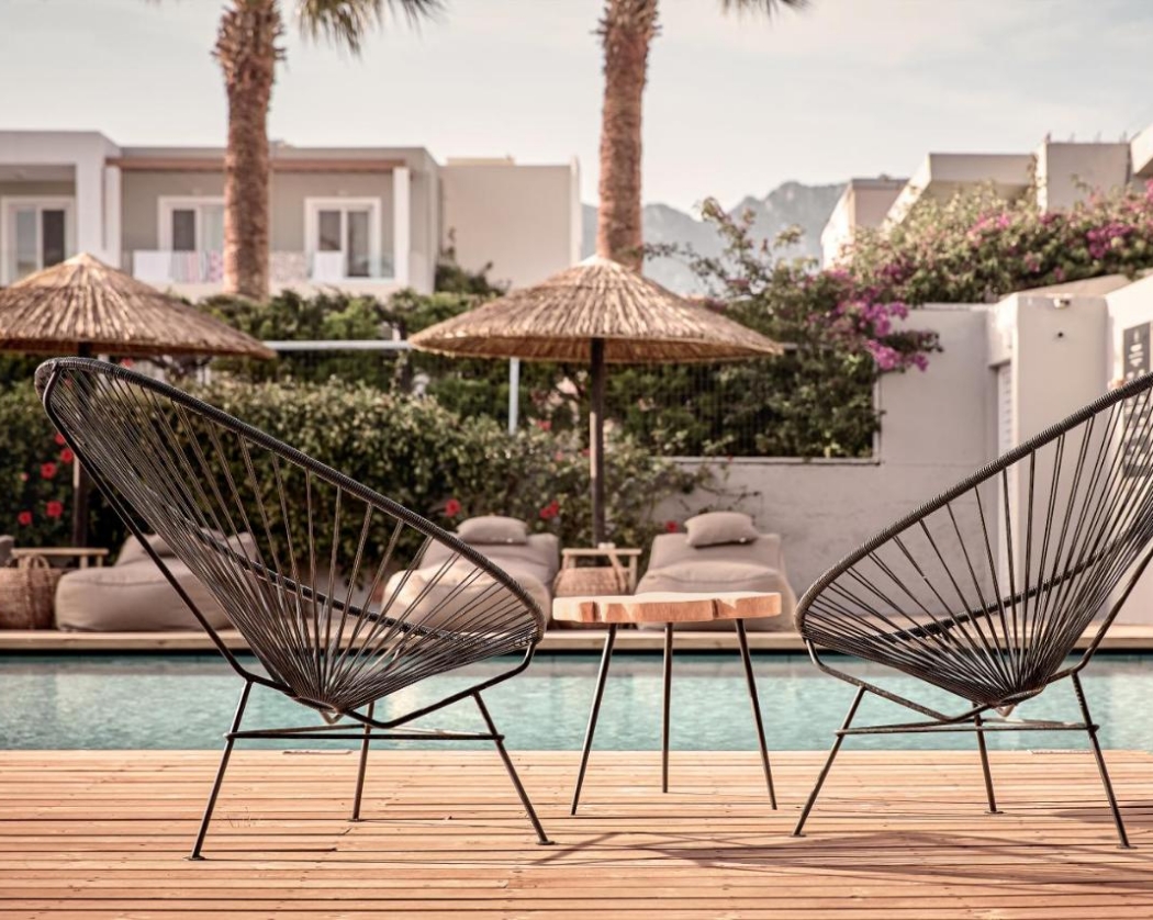 Zwei moderne schwarze Drahtstühle und ein kleiner Holztisch stehen auf einer Holzterrasse vor einem Pool, im Hintergrund stilvolle weiße Hotelgebäude, Palmen, Strohdächer und blühende Bougainvillea.