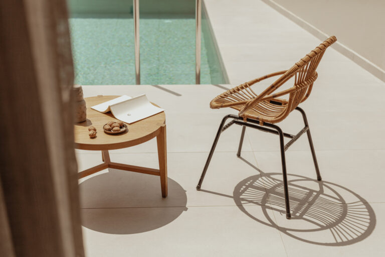 Ein stilvoller Stuhl steht neben einem runden Holztisch mit einem offenen Notizbuch, einer Tonvase und einer Schale mit Gebäck, direkt am Rand eines privaten Pools in der warmen Nachmittagssonne.