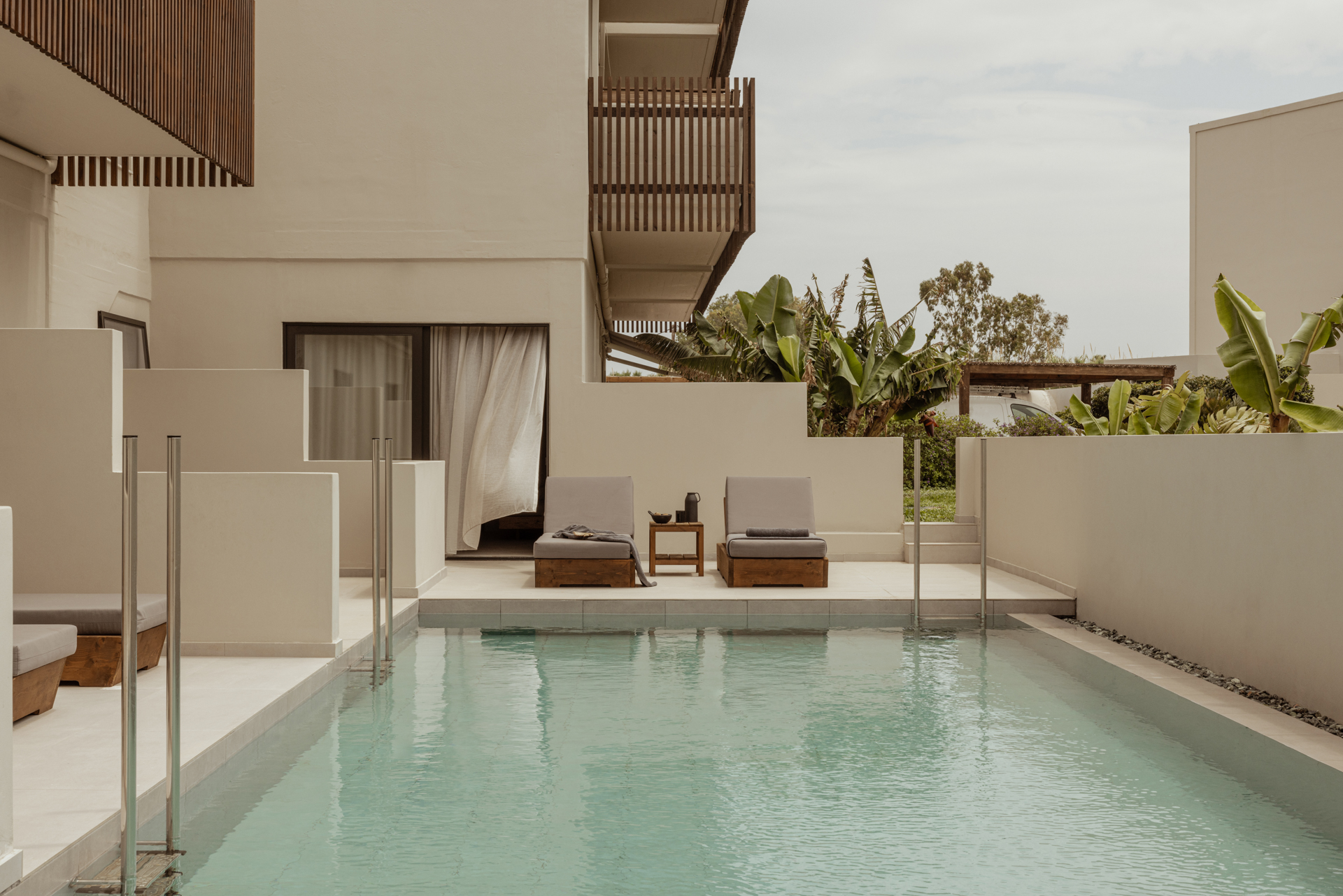 Moderner Hotelbereich mit direktem Poolzugang, zwei grauen Liegen auf einer privaten Terrasse, umgeben von weißen Wänden und tropischer Bepflanzung im Hintergrund, unterhalb von Holzbalkonen.