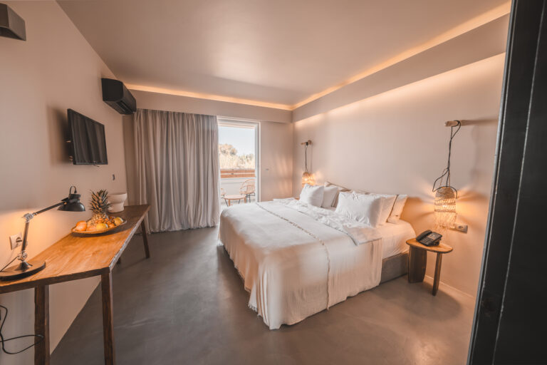 Modern eingerichtetes Hotelzimmer mit großem Doppelbett, Wandbeleuchtung, Schreibtisch mit Obstschale und Schreibtischlampe, sowie Zugang zu einem Balkon mit zwei Stühlen und Ausblick auf Bäume im Sonnenlicht.