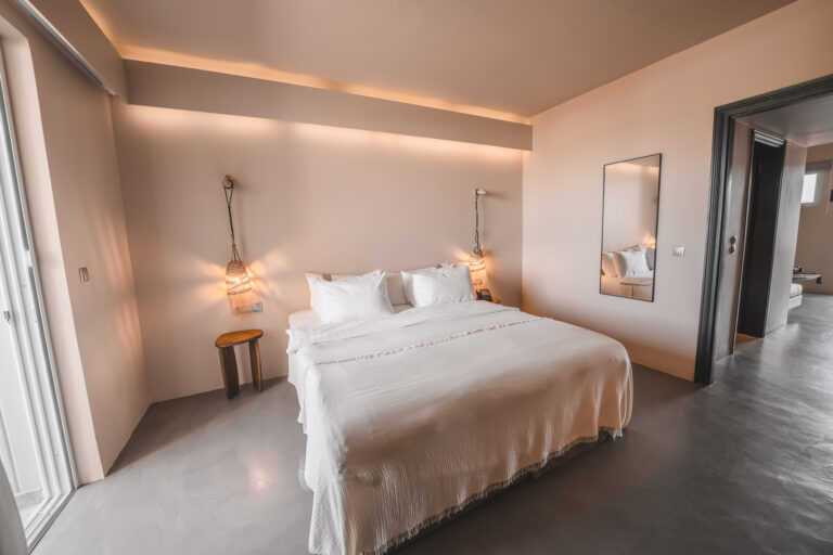Modernes, minimalistisches Schlafzimmer mit einem großen Doppelbett, zwei hängenden Rattanlampen, einem schlichten Wandspiegel und Zugang zu einem angrenzenden Raum mit Sicht auf ein weiteres Bett.