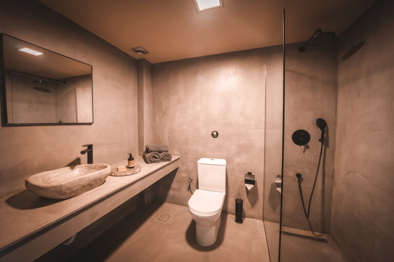 Modernes Badezimmer in warmen Beigetönen mit offener Dusche, schwarzer Regenbrause, einem weißen WC und einem breiten Waschtisch mit natursteinartigem Waschbecken, schwarzer Armatur und gerollten Handtüchern.