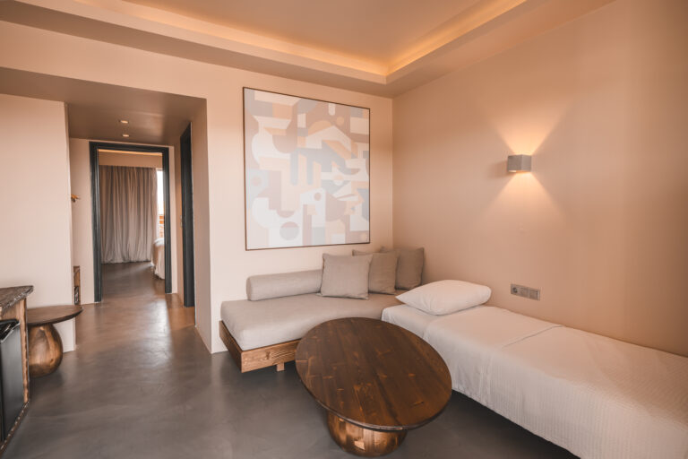 Helles, modern eingerichtetes Hotelzimmer mit zwei Tagesbetten, mehreren Kissen, einem ovalen Holztisch und abstrakter Wandkunst; durch die offene Tür ist ein weiteres Schlafzimmer mit Doppelbett sichtbar.