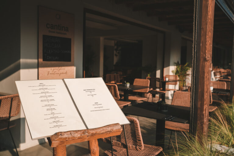 Einladender Außenbereich eines griechischen Restaurants mit gedeckten Tischen und einem aufgeschlagenen Menü auf Holzständer im Vordergrund, beleuchtet vom warmen Sonnenlicht.