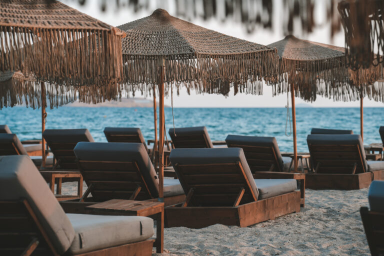 Reihe komfortabler Sonnenliegen mit geflochtenen Schirmen am Sandstrand eines stilvollen Strandresorts mit Blick auf das ruhige, blaue Meer bei Sonnenuntergang.