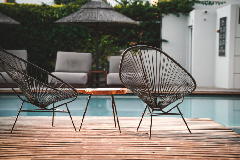 Zwei elegante Lounge-Stühle mit filigranem Metallgestell und kleinem Holztisch auf einer Holzterrasse am Pool eines stilvollen Resorts, im Hintergrund Sonnenliegen und grüne Hecken.