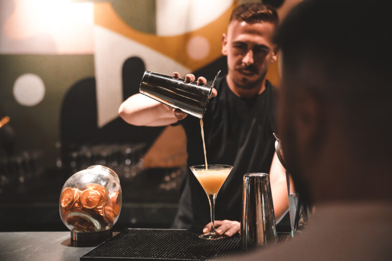 Barkeeper beim stilvollen Einschenken eines Cocktails in ein Martiniglas an einer eleganten Hotelbar mit künstlerischer Wandgestaltung und gedämpfter Lichtstimmung.
