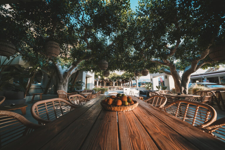 Langer Holztisch mit Korbstühlen unter schattenspendenden Bäumen in einem offenen, mediterranen Hotelrestaurant; dekorative Körbe hängen in den Ästen, eine Obstschale steht auf dem Tisch.