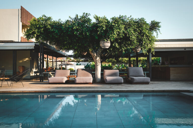 Moderne Poolterrasse mit breiten, gepolsterten Sonnenliegen unter einem schattigen Baum mit dekorativen Lampen, im Hintergrund Bar- und Sitzbereich eines Resorts.