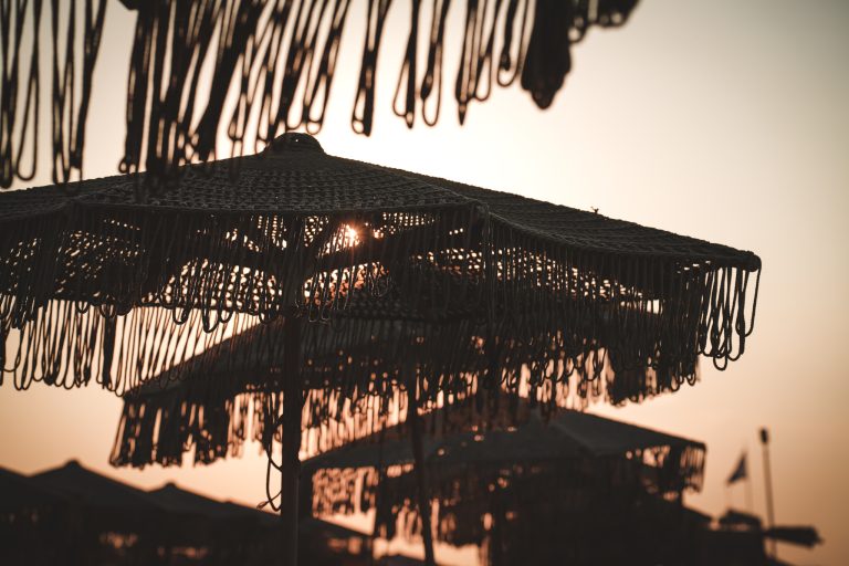 Makramee-Sonnenschirm am Strand bei Sonnenuntergang, mit warmem Licht, das durch das geflochtene Muster scheint.