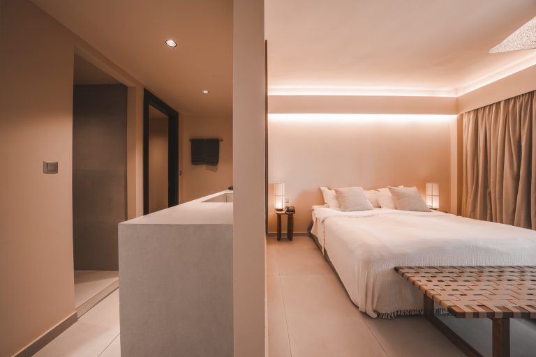 Modern eingerichtetes Hotelzimmer mit einem großen Doppelbett, warmen indirekten Lichtquellen, minimalistischem Badezimmer und gedeckten Farbtönen in Beige und Sand.