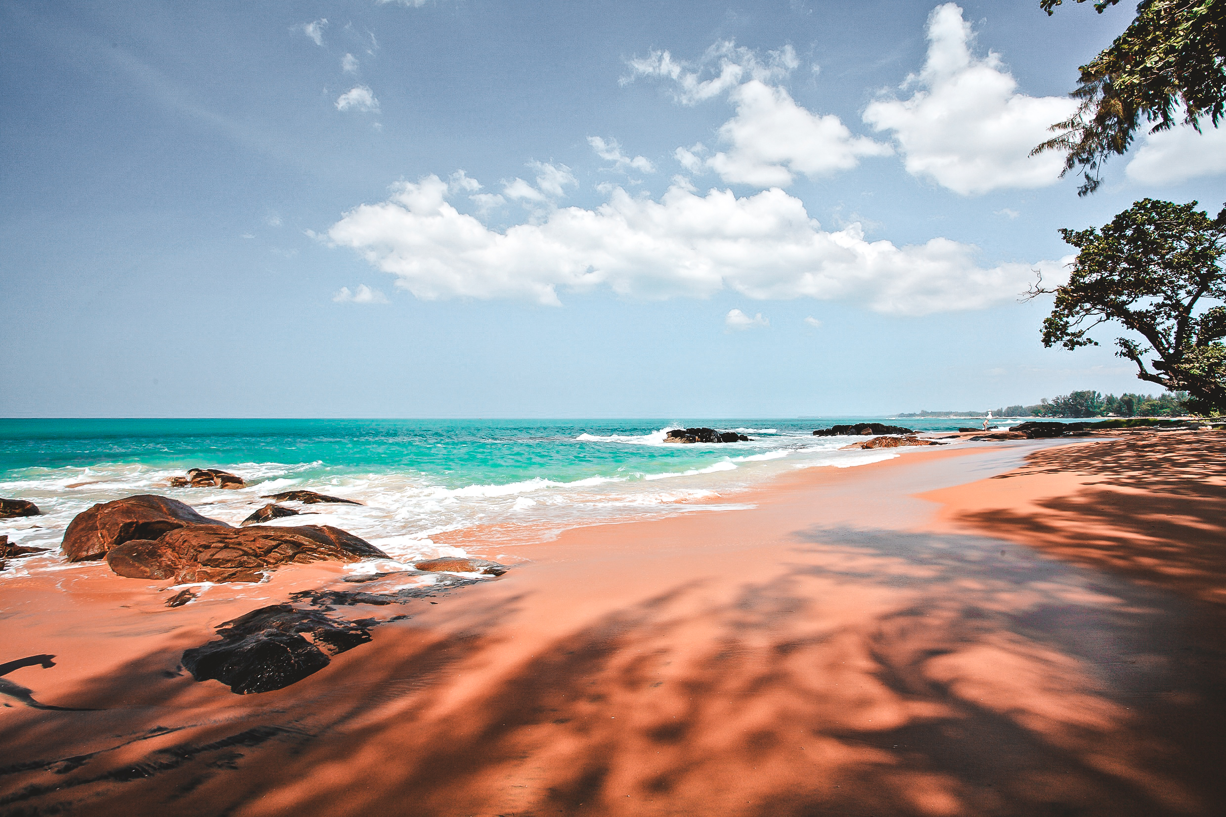 Roter Sandstrand mit großen Felsen, türkisfarbenem Meer und blauem Himmel mit weißen Wolken, Schatten von Bäumen auf dem Sand.