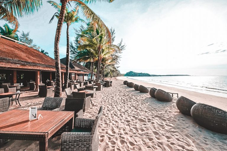 Ruhige Strandbar mit Tischen und Stühlen aus Rattan unter Palmen, hellem Sandstrand mit Sitzkörben am Wasser und ruhigem Meer im Hintergrund.