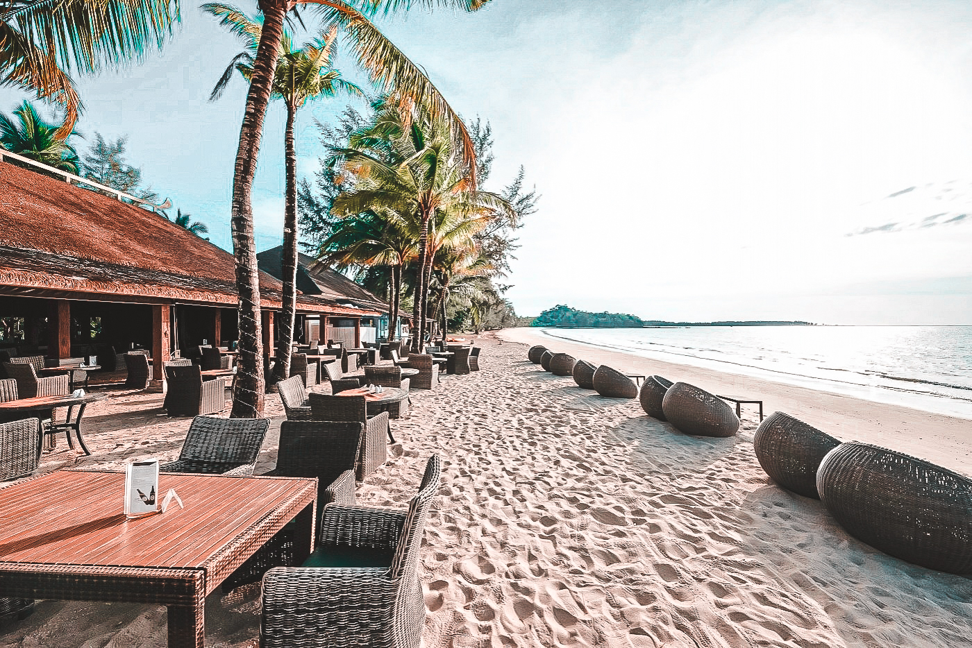 Ruhige Strandbar mit Tischen und Stühlen aus Rattan unter Palmen, hellem Sandstrand mit Sitzkörben am Wasser und ruhigem Meer im Hintergrund.