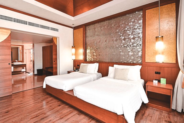 Helles Hotelzimmer mit zwei Einzelbetten, Holzfußboden, eleganter Wanddekoration und zwei hängenden Lampen neben den Betten, Blick ins angrenzende Badezimmer.