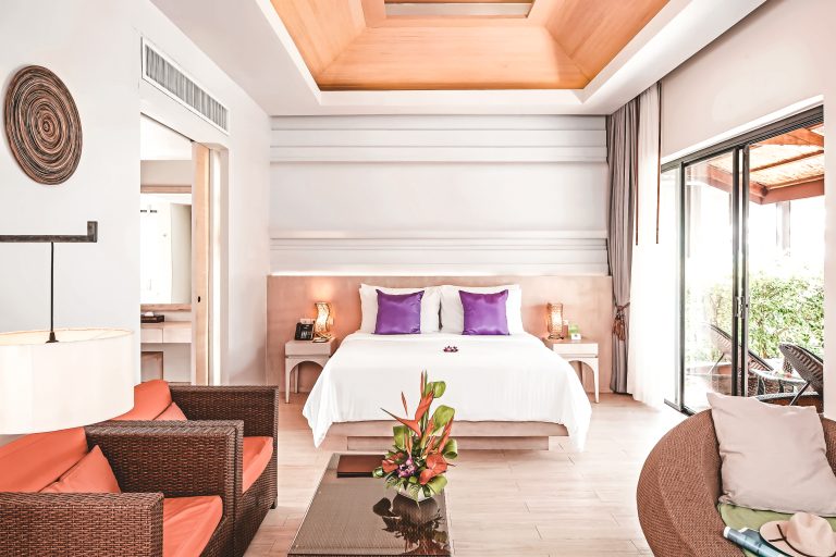 Helle, moderne Hotelzimmer-Suite mit Doppelbett, lila Kissen, orangefarbenem Sofa, Rattan-Sessel, Blumendekoration und großem Schiebefenster zum Garten.
