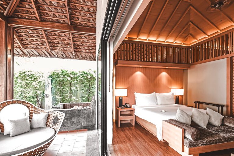 Moderner Hotelzimmer-Balkon mit Rattan-Lounge und Holzbett, weiße Bettwäsche, Holzboden, tropische Pflanzen draußen, natürliche Beleuchtung.