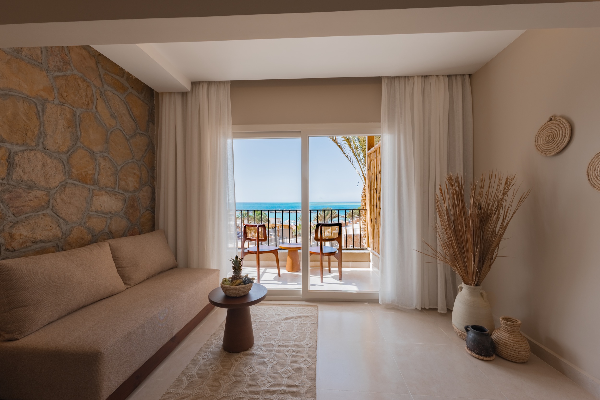 Gemütlicher Wohnbereich mit braunem Sofa, kleinem Tisch mit Obstkorb, großen Fenster mit Balkon und Meerblick, dekorativen Körben und Trockengras.