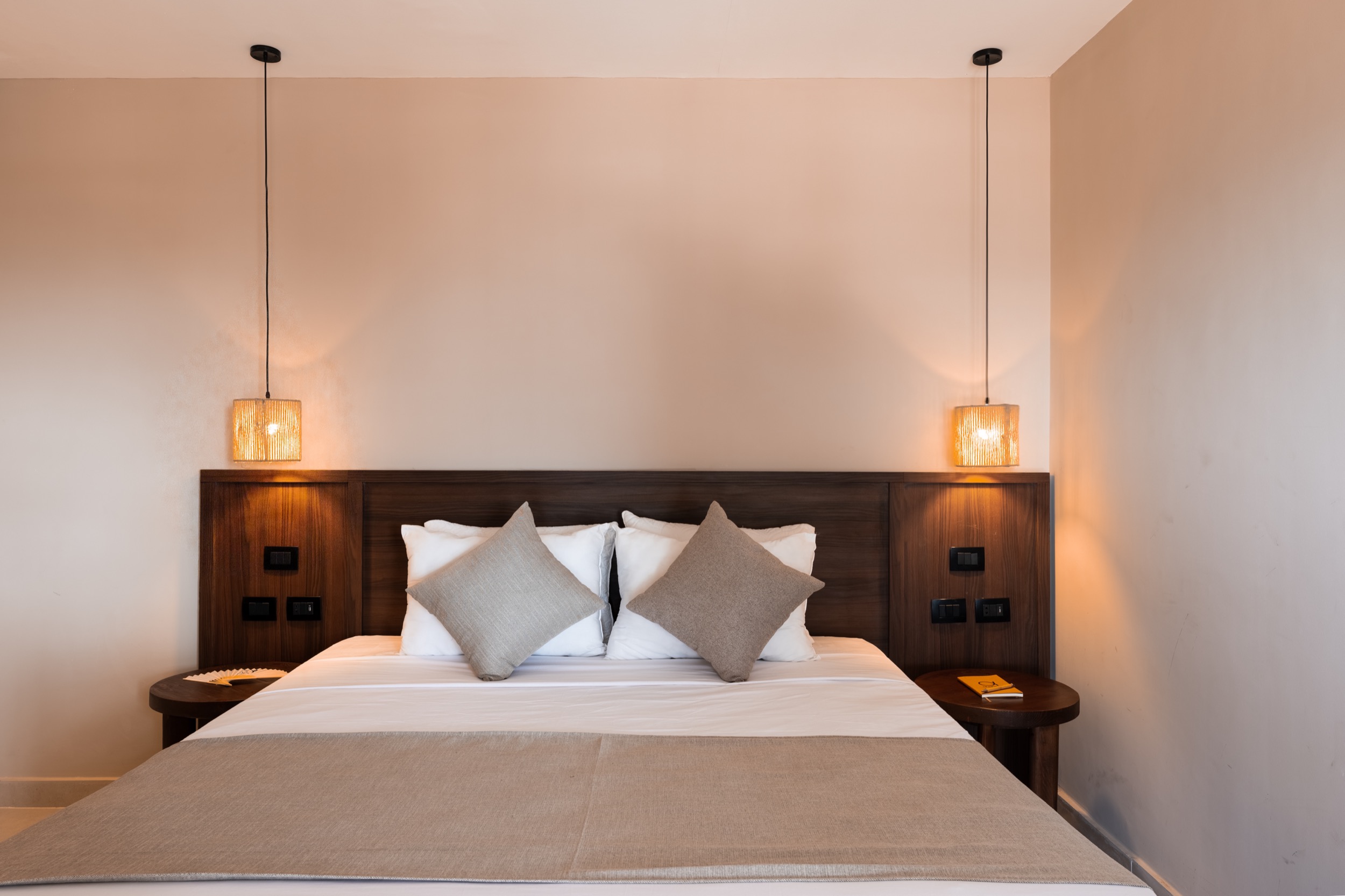 Doppelbett mit Holzrahmen, zwei Hängelampen, neutralen Kissen und Nachttischen in einem Hotelzimmer.