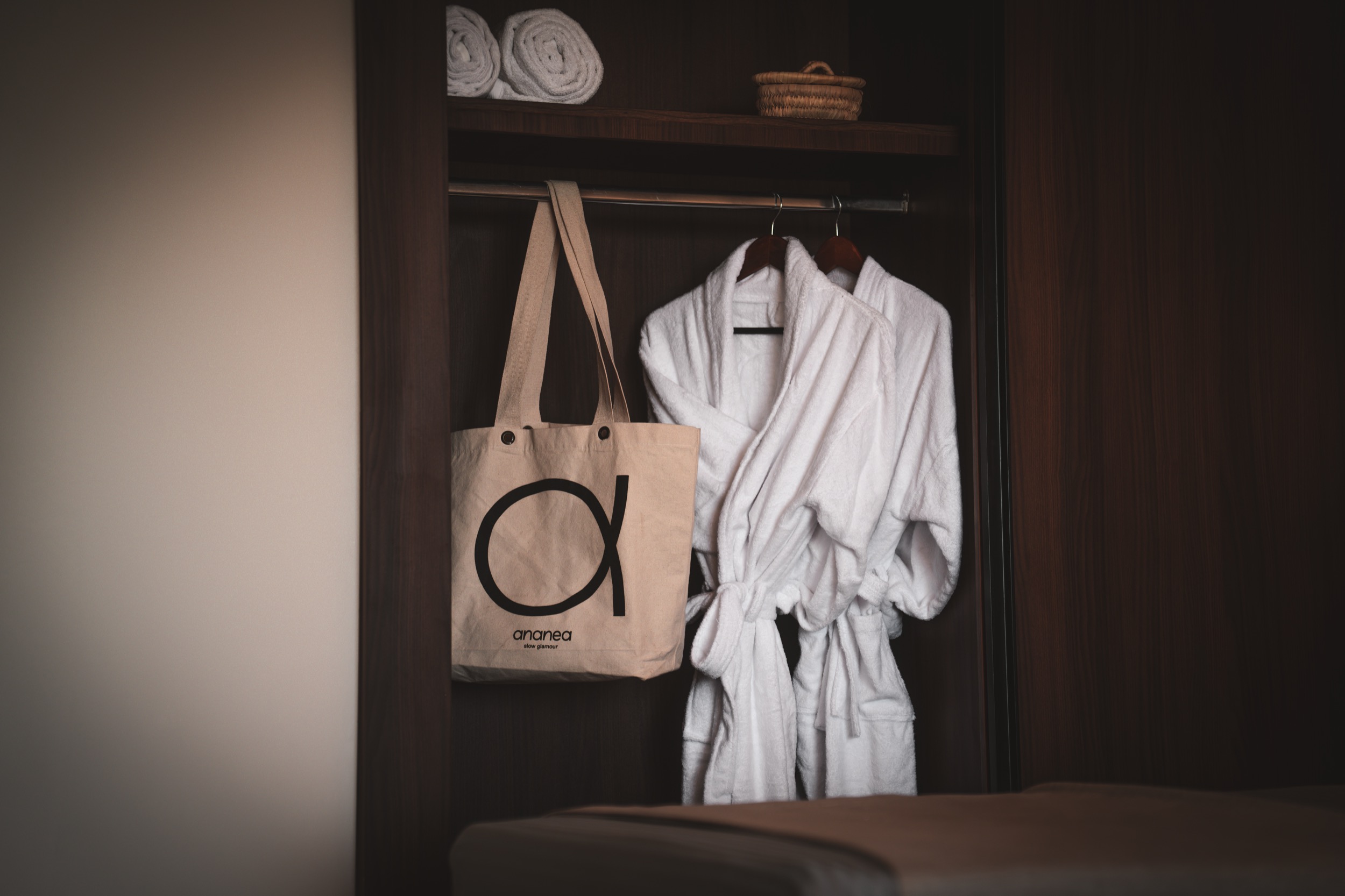 Zwei weiße Bademäntel hängen in einem dunklen Holzschrank, daneben eine Tasche mit Logo "aranea slow glamour".