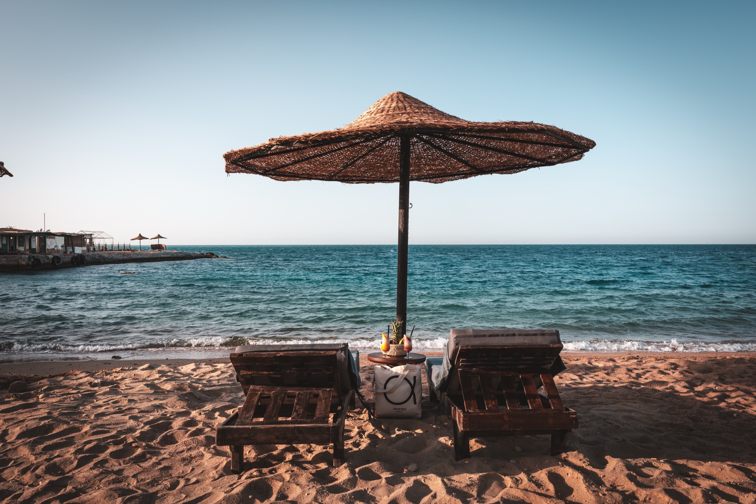 Strand mit zwei Holzliegen unter einem Sonnenschirm aus geflochtenem Material, im Hintergrund blaues Meer und bewölkter Himmel.