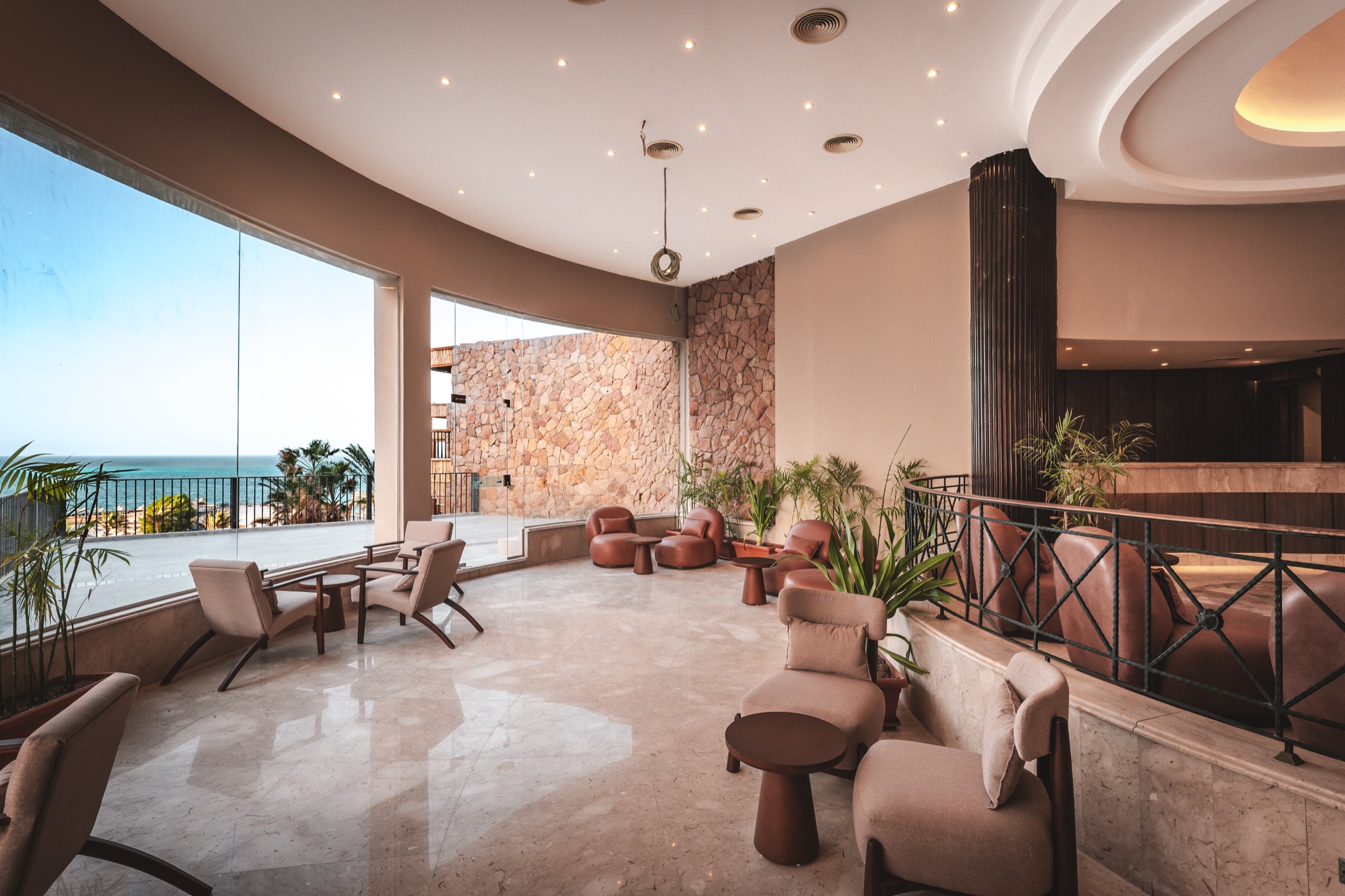 Moderne Lounge mit Sesseln, Tischen, großen Fenstern und Meerblick, warme Erdtöne, Natursteinwand, helle Atmosphäre.
