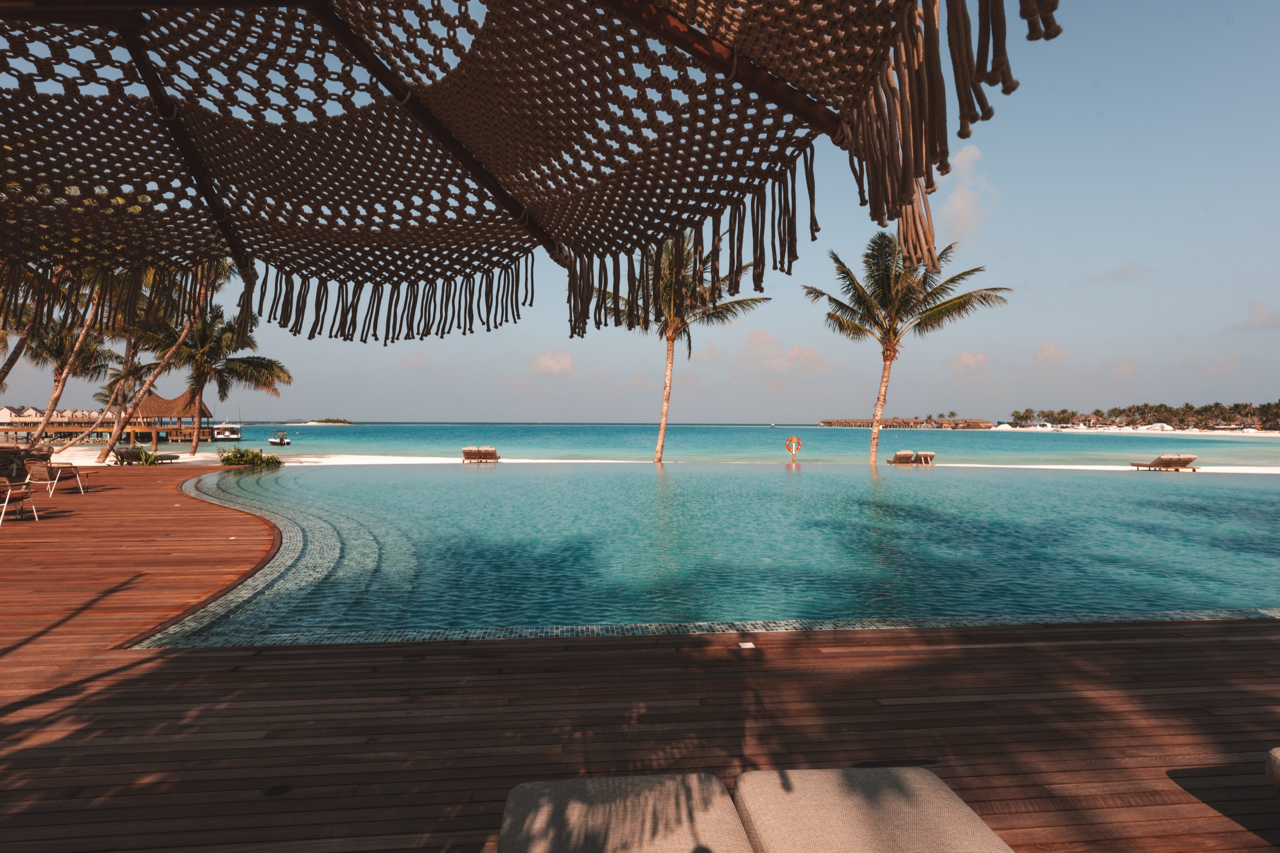 Blick auf einen Pool mit klarem Wasser, umgeben von Holzdeck und einem Sonnenschirm aus geflochtenem Material. Im Hintergrund ist das türkisfarbene Meer und der Himmel zu sehen.“