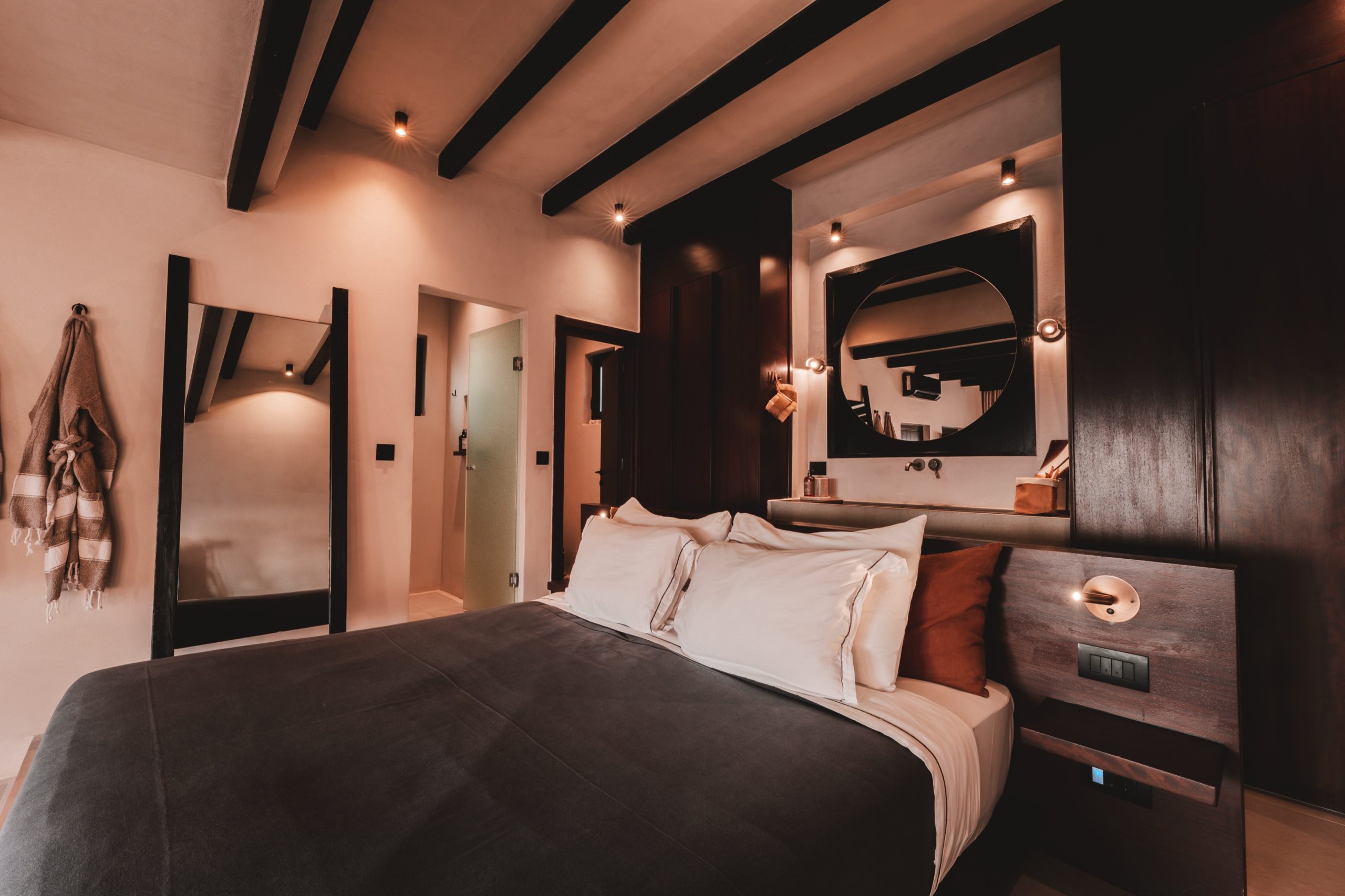 Modernes Hotelzimmer mit Doppelbett, dunklen Holzelementen, Spiegel, Badzugang und minimalistischem Design.