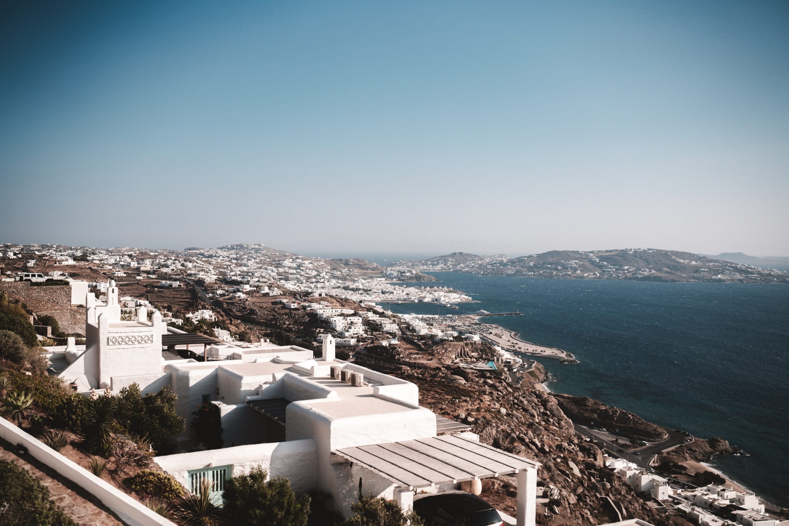Blick auf eine Küstenstadt mit weißen Häusern, Hügeln und Meer, vermutlich auf einer griechischen Insel.