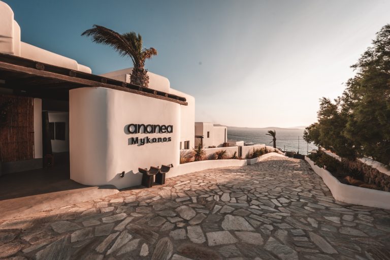 Eingang des Hotels Ananea Mykonos mit weißer Fassade, Palmen, Kopfsteinpflasterweg und Meerblick bei Sonnenuntergang.