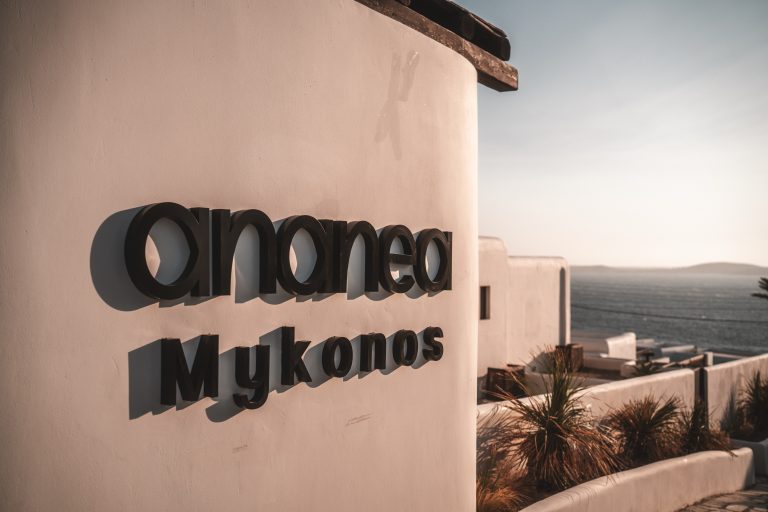 Hotellogo „ananea Mykonos“ an weißer Fassade mit Blick auf das Meer und umliegende, helle Gebäude bei Sonnenuntergang.