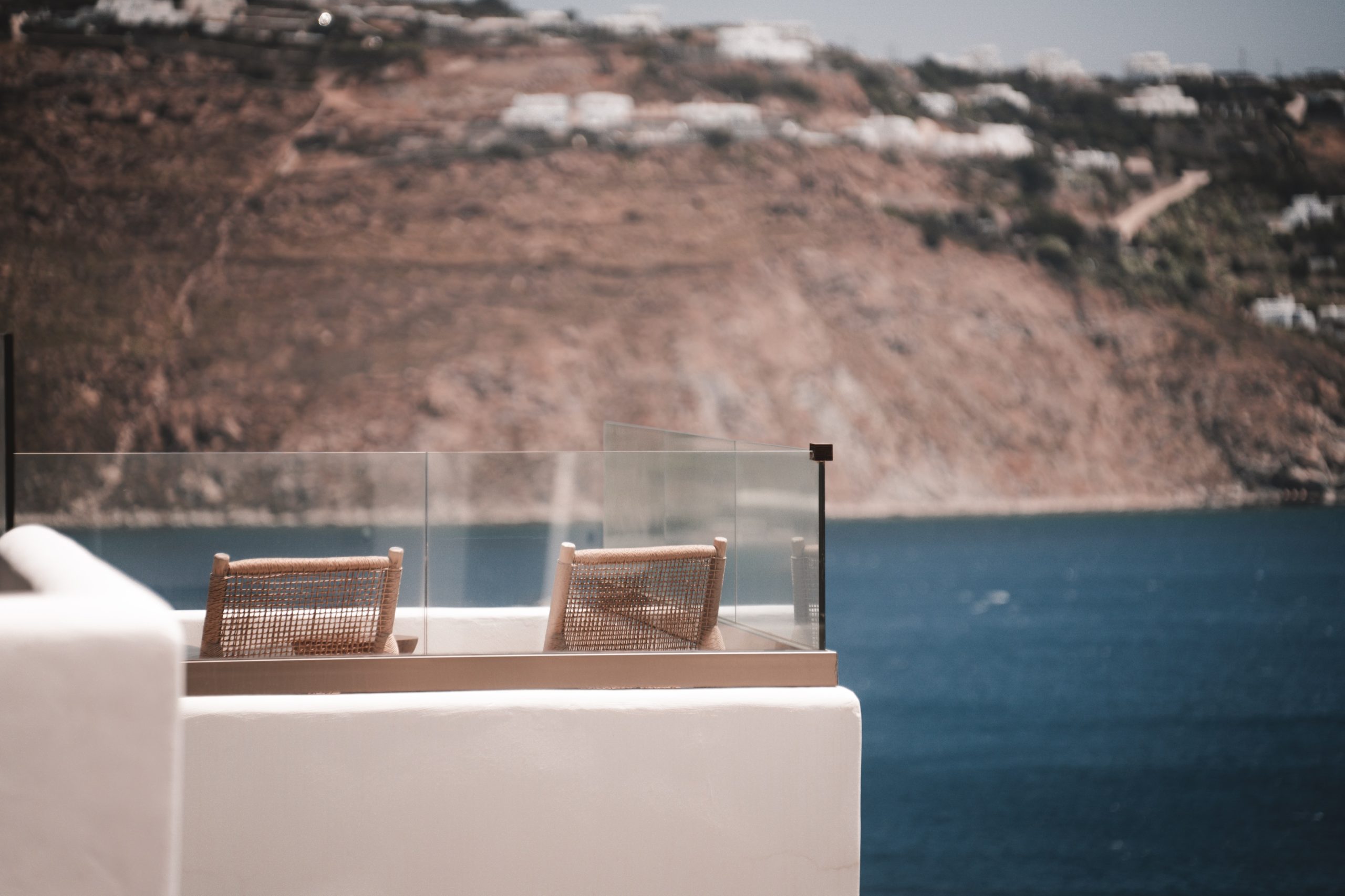 Zwei geflochtene Holzstühle stehen auf einer Terrasse mit Glasbrüstung, dahinter Blick aufs Meer und felsige Küste.