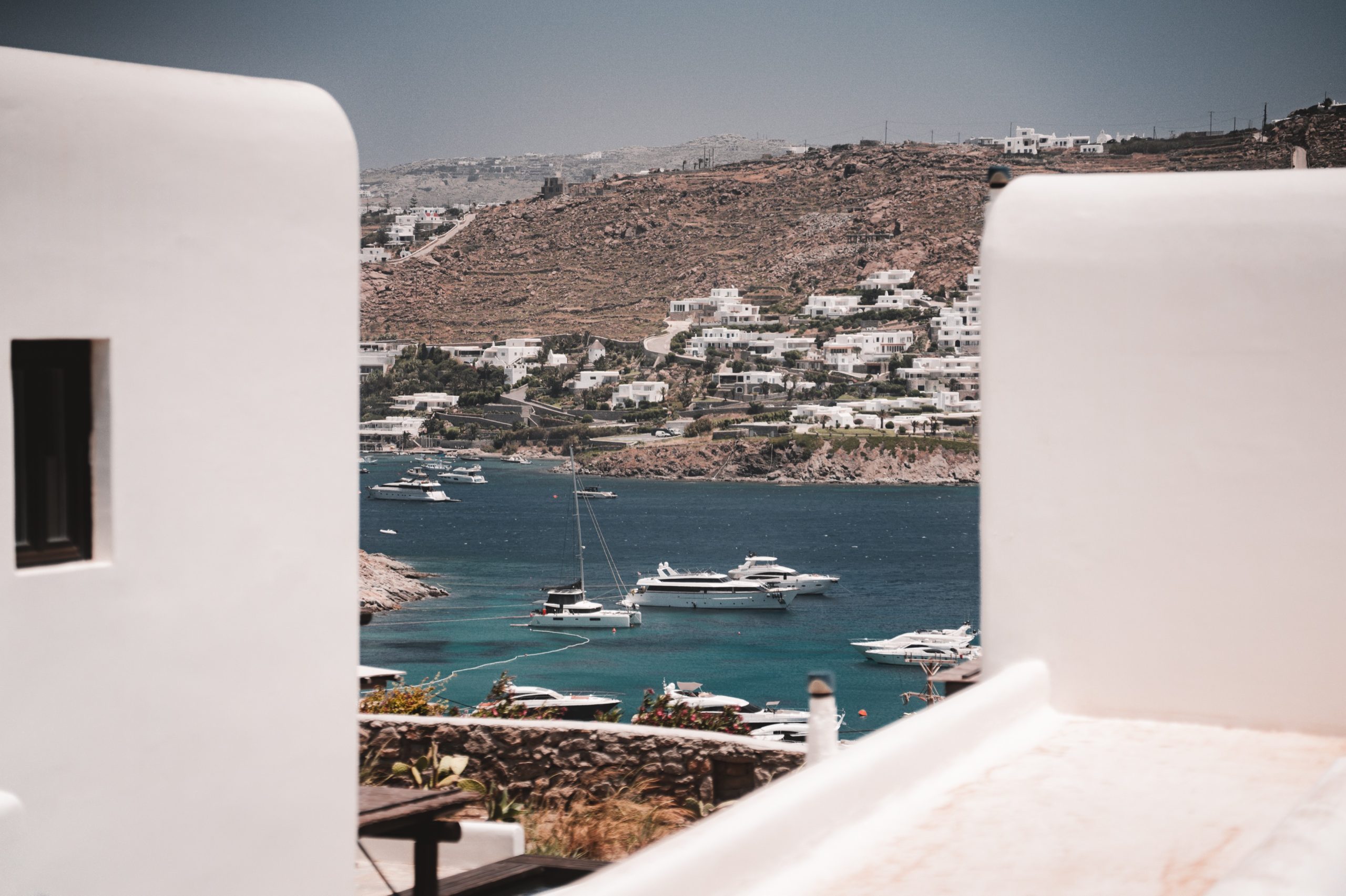 Blick durch weiße Häuser auf eine Bucht mit Yachten und Segelbooten vor der Küste einer mediterranen Hügellandschaft.