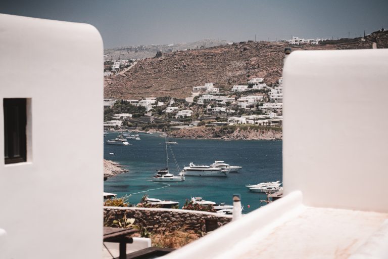 Blick zwischen weißen Gebäuden auf Yachten im türkisblauen Meer und weiße Häuser an einem felsigen Hügel auf Mykonos.