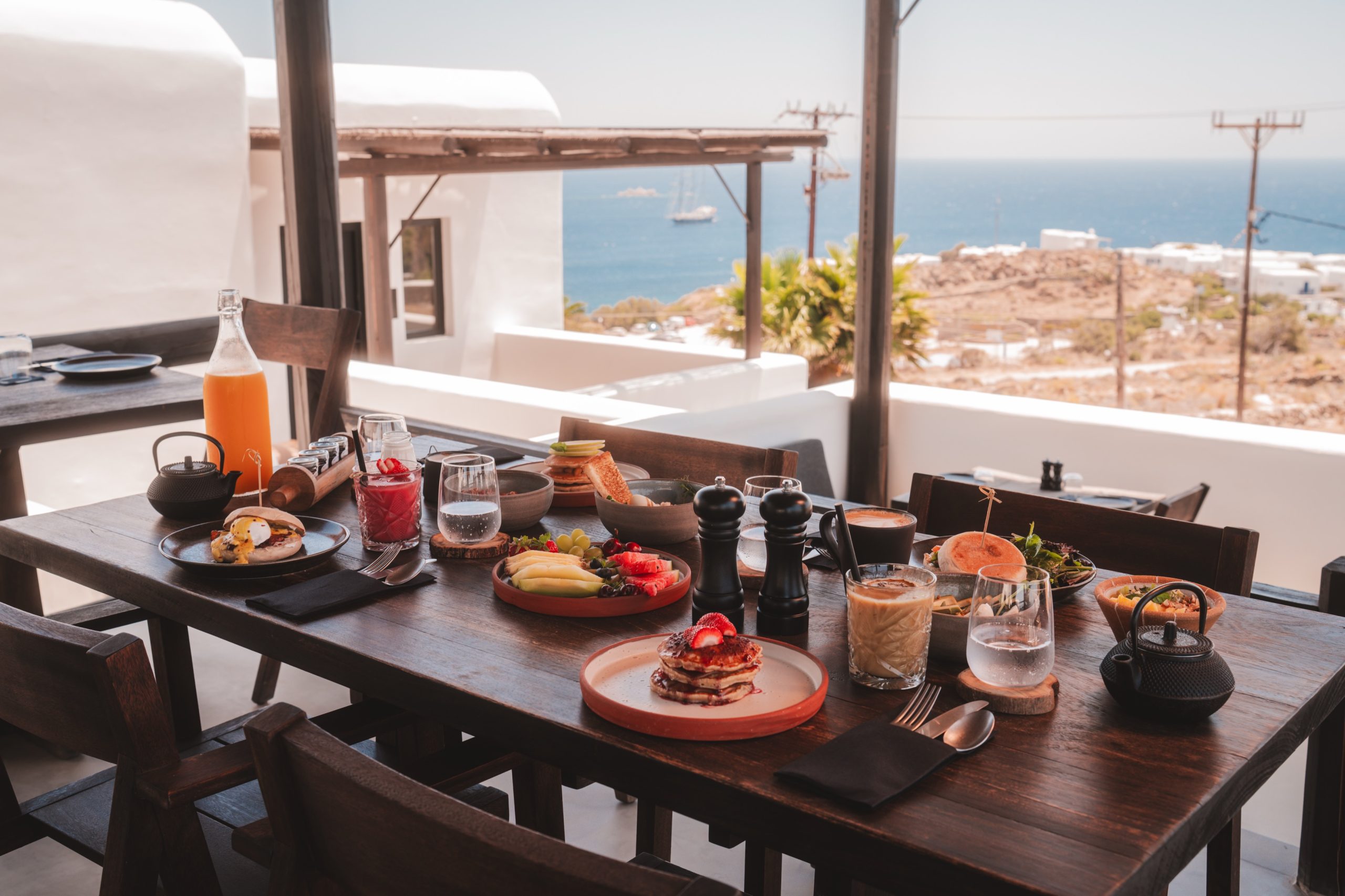 Frühstückstisch mit frischem Obst, Pancakes, Eiern und Kaffee auf einer Terrasse mit Meerblick auf Mykonos.