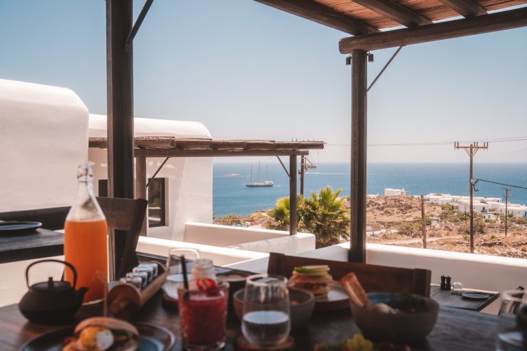 Frühstück mit Meerblick auf Mykonos, frische Getränke und Speisen auf der Terrasse mit Blick auf die Ägäis und Segelschiff.