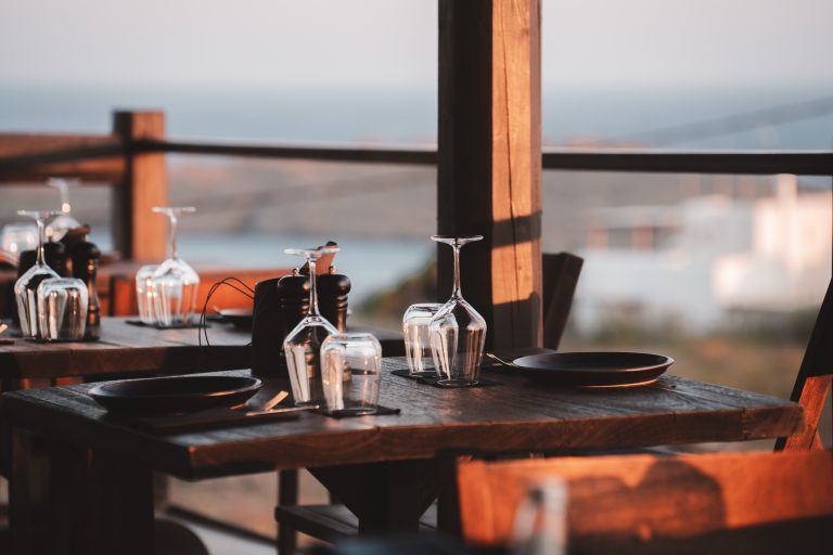 Gedeckter Holztisch in einem Restaurant mit Blick aufs Meer, Gläser und Teller spiegeln das warme Sonnenlicht wider.