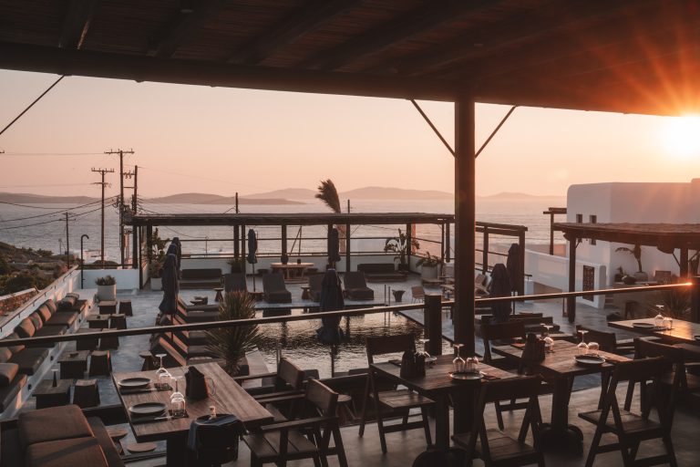 Sonnenterrasse eines Restaurants mit Meerblick bei Sonnenuntergang, gedeckte Holztische und gemütliche Sitzbereiche.