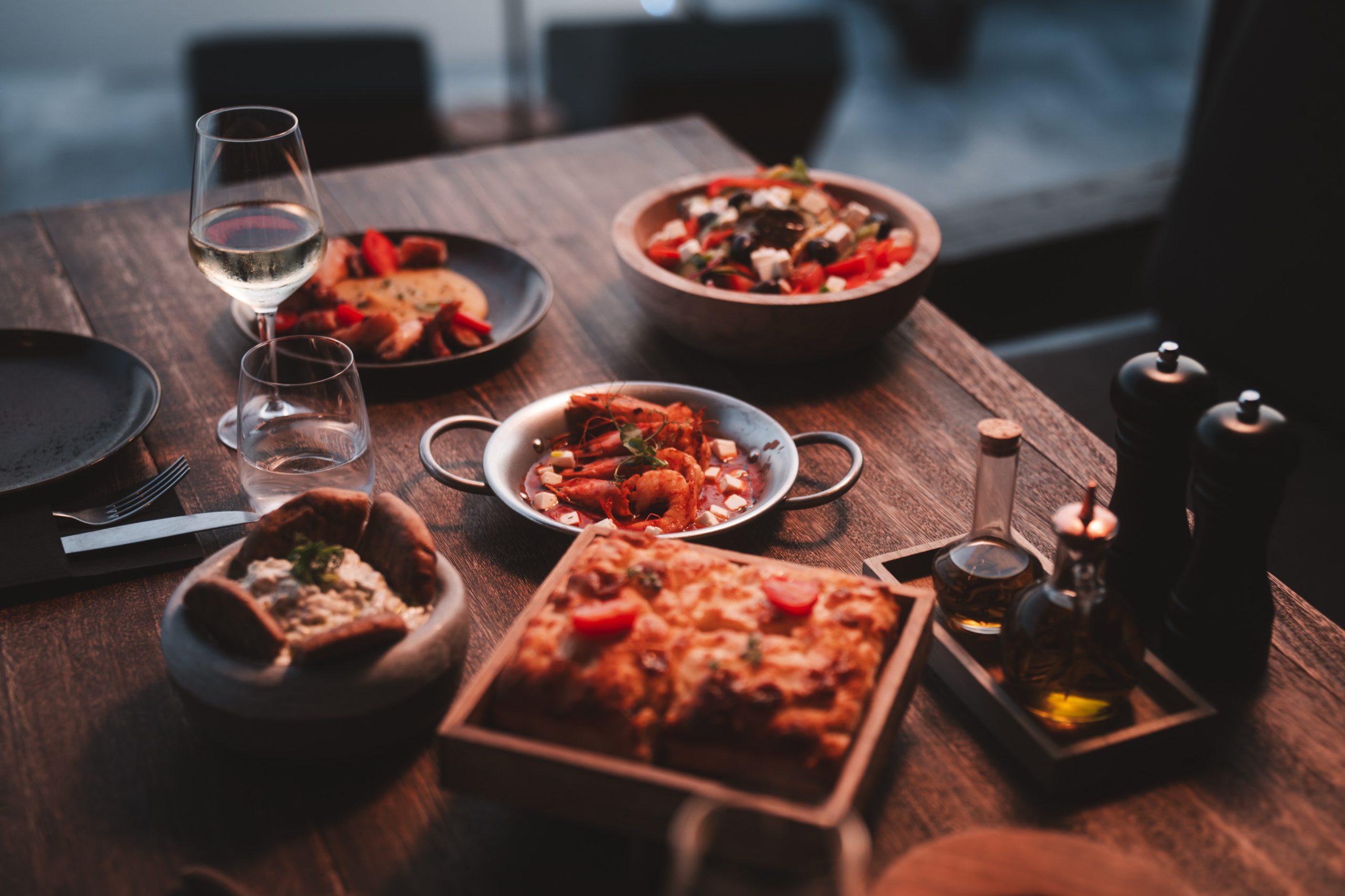 Mediterraner Abendtisch mit Wein, Brot, Meeresfrüchten, Dip, Salat und Olivenöl auf rustikalem Holztisch.
