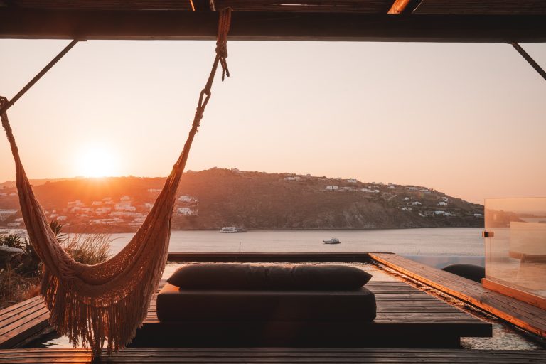 Hängematte und Sonnenliege auf Holzterrasse mit Blick auf die Ägäis und Hügel von Mykonos bei Sonnenuntergang.