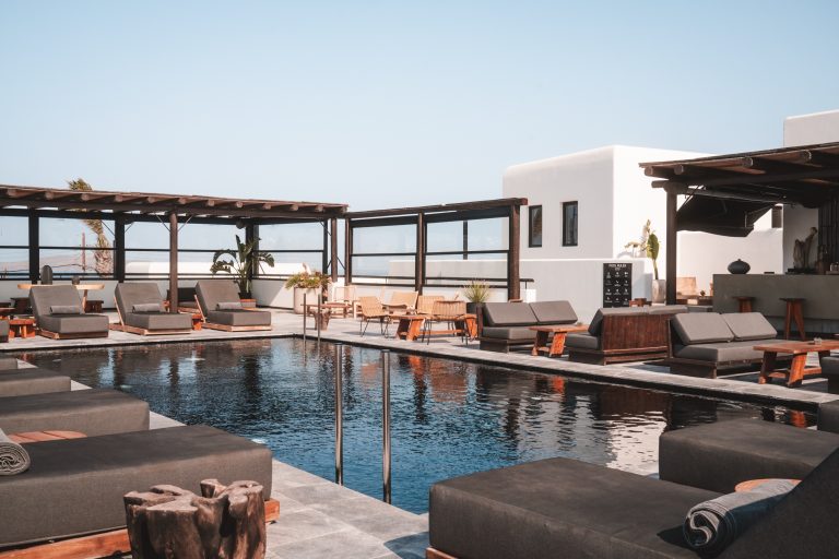 Moderner Poolbereich mit eleganten grauen Sonnenliegen, Holzmöbeln und weißem Gebäude im Hintergrund auf Mykonos.