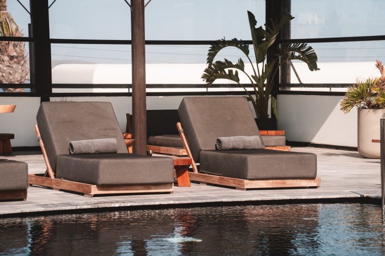 Zwei moderne graue Sonnenliegen mit gerollten Handtüchern stehen am Pool eines stilvollen Resorts mit Pflanzen im Hintergrund.