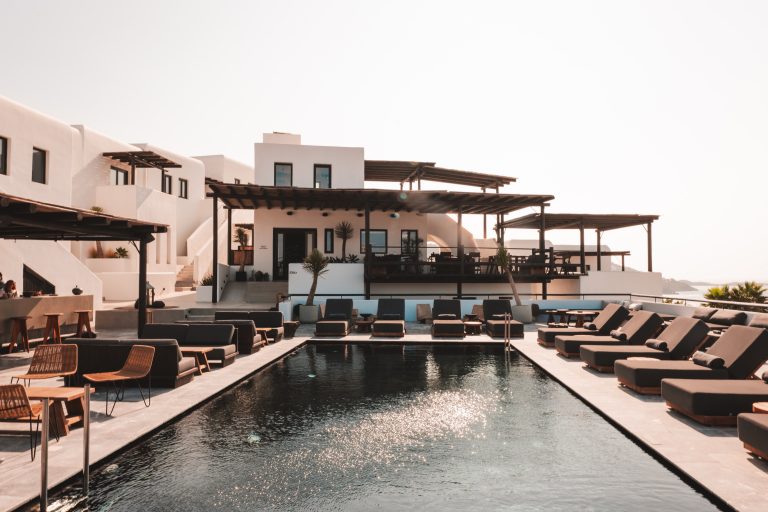 Moderner Hotelpool auf Mykonos mit Sonnenliegen, weißen Gebäuden im Kykladenstil und schattiger Barterrasse.