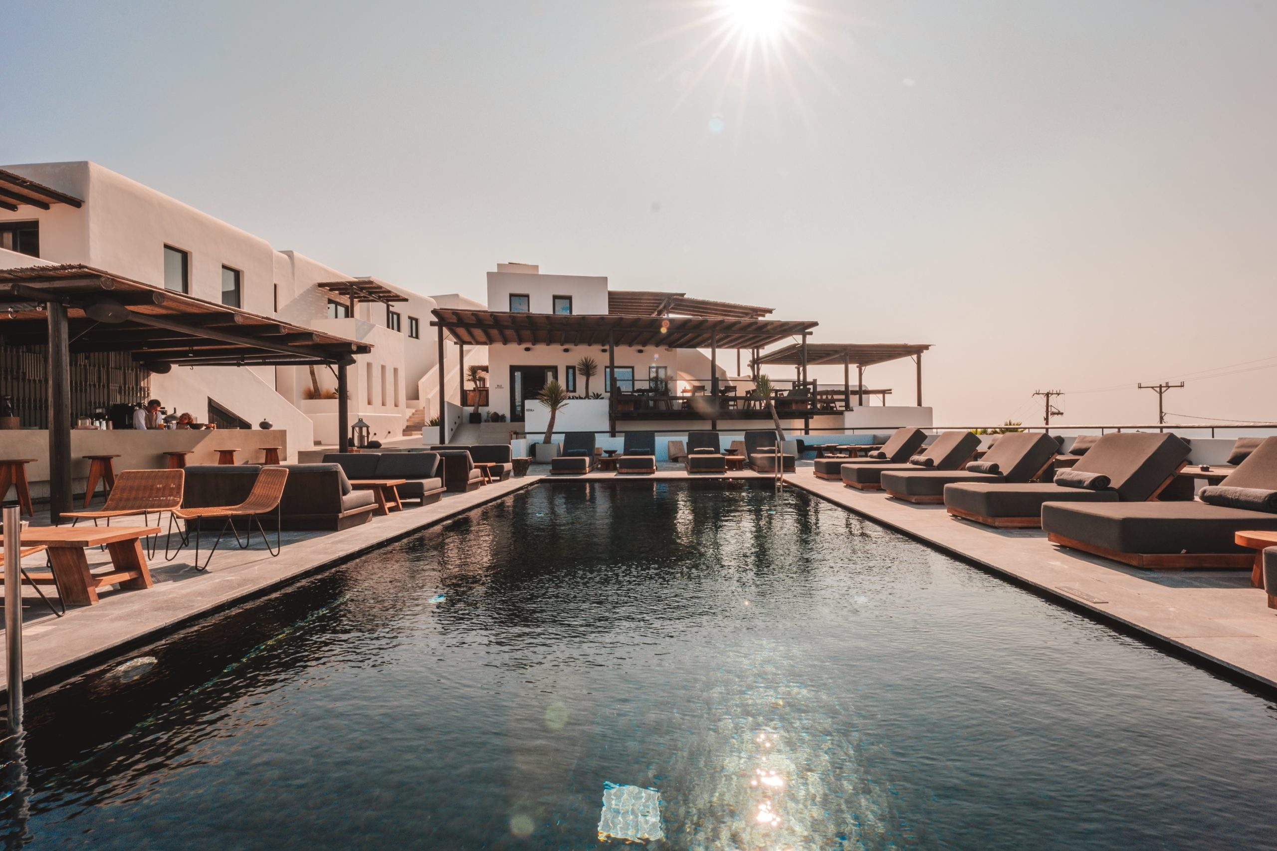 Moderner Hotelpool mit Sonnenliegen, Barbereich und weißen Gebäuden unter strahlender Sonne und klarem Himmel.
