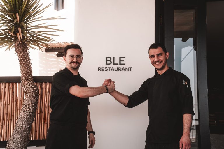 Zwei Köche in schwarzer Kleidung posieren lachend vor dem BLE Restaurant auf Mykonos und schütteln sich die Hände.