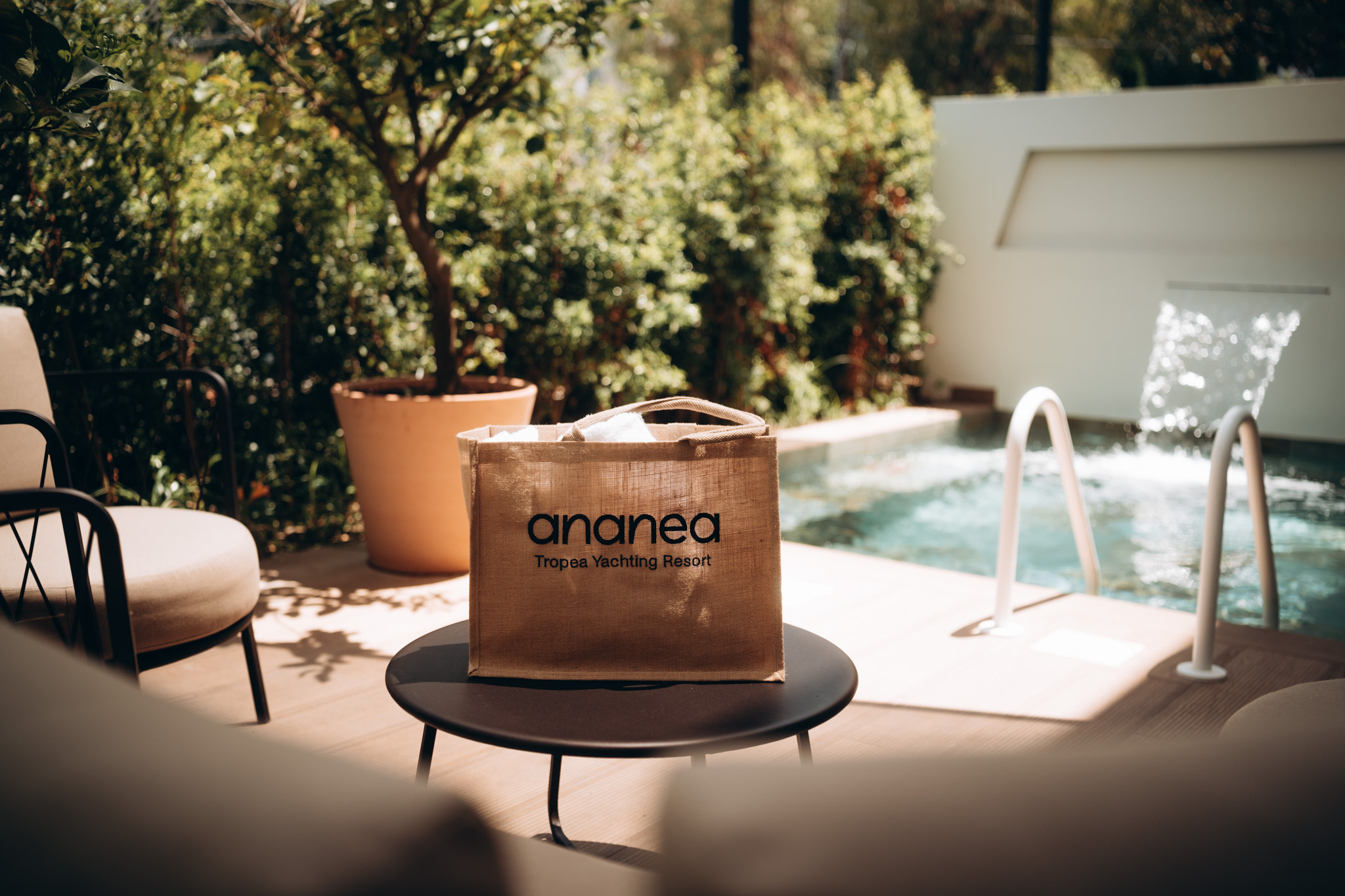 Jutetasche mit dem Aufdruck "ananea Tropea Yachting Resort" steht auf einem schwarzen Tisch neben einem Pool mit Wasserfontäne, umgeben von Pflanzen und Outdoor-Möbeln, bei sonnigem Wetter.