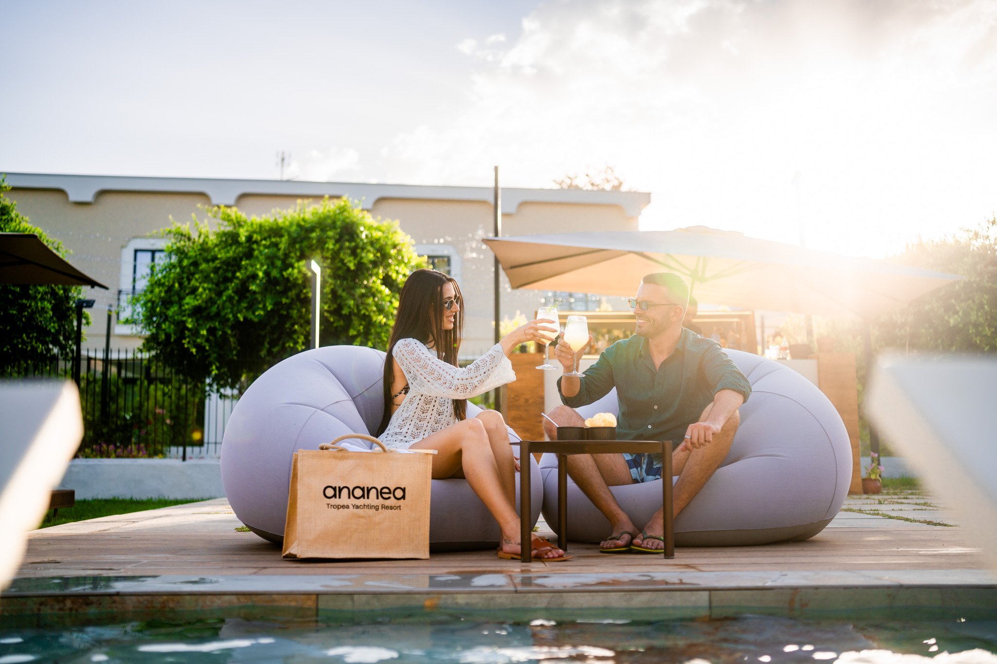 Junges Paar sitzt entspannt auf grauem Loungsesseln am Pool, stößt mit Drinks an, Einkaufstasche mit "ananea Tropea Yachting Resort" neben ihnen.