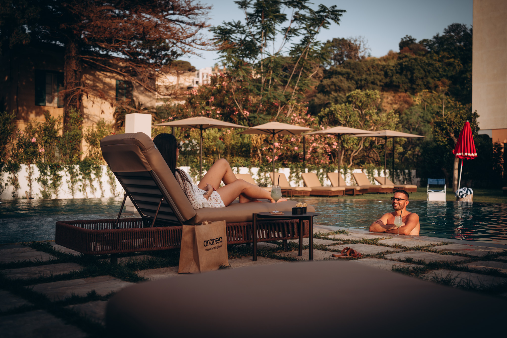 Frau entspannt auf Liegestuhl am Poolrand, Mann genießt Getränk im Pool, umgeben von Sonnenschirmen und Liegestühlen, grüner Garten mit Bäumen und Blumen, spätnachmittägliches warmes Licht.