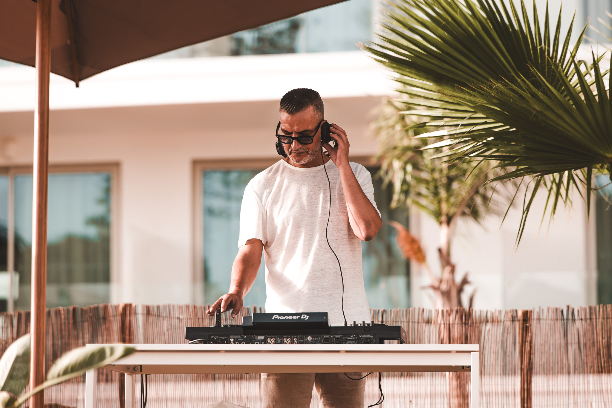 „DJ mit Kopfhörern legt im Freien an einem Mischpult auf, umgeben von Palmen und entspannter Urlaubsatmosphäre.“